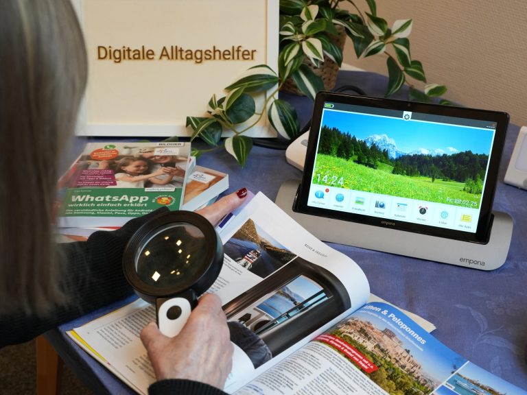 Foto eines Aufbausettings der Digitalen Alltagshelfer. Hände einer Frau mit Lupe, Seniorentablet, Ratgeberbuch