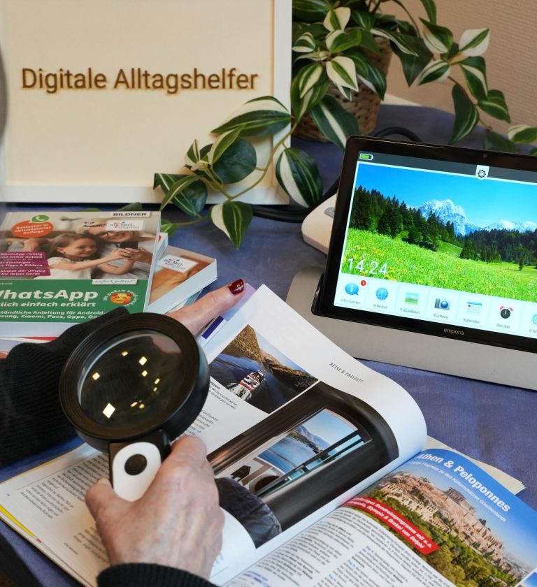 Foto eines Aufbausettings der Digitalen Alltagshelfer. Hände einer Frau mit Lupe, Seniorentablet, Ratgeberbuch